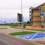 Miejsca parkingowe prostopadłe do ulicy, w tym jedno miejsce dla osób z niepełnosprawnościami pomalowane na niebiesko. W tle budowa obiektu.