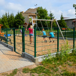Zdjęcie przedstawia ogrodzony plac zabaw, na którym bawią się dzieci