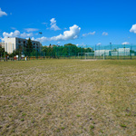 Na zdjęciu ogrodzone boisko trawiaste oraz siłownia pod chmurką