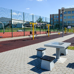 Zdjęcie przedstawia urządzenia siłowni zewnętrznej,  z tyłu boisko. Na pierwszym planie betonowy stół do gry w szachy wraz z ławeczkami.