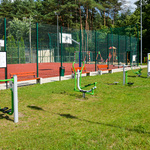 Zdjęcie przedstawia ogrodzone boisko wielofunkcyjne, siłownię oraz plac zabaw z ławeczkami