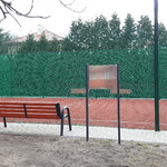 Boisko pod kasztanowcem ogrodzone piłkochwytami, przed boiskiem ustawiona jest ławka