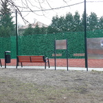 Boisko pod kasztanowcem ogrodzone piłkochwytami, przed boiskiem ustawiona jest ławka
