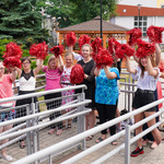 Grupa uśmiechniętych dziewcząt stoi na podjeździe przed szkołą. W podniesionych do góry dłoniach trzymają czerwone pompony.