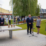 Zdjęcie terenu zielonego przed szkołą. Prezydent przemawia do mikrofonu. Po prawej stoi kilka osób, po lewej stół do pingponga na którym stoją statuetki w kształcie serc. Za nim stoi kilka osób.