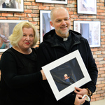 Ksiądz pozuje do zdjęcia z kobietą o blond włosach. W dłoniach trzyma ramę z fotografią z wystawy.