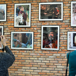 Dwie kobiety stoją przodem do ceglanej ściany, na której wiszą duże fotografie w ramach. Robią zdjęcia telefonami.