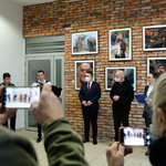 Kilka osób stoi przy ceglanej ścianie, na której wiszą fotografie. Kilka osób robi zdjęcia.