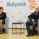 Ksiądz, który jest autorem zdjęć siedzi na fotelu. Wskazuje dłonią w stronę prowadzącej spotkanie, która siedzi na fotelu obok. W tle jasna ścianka reklamowa z dużym logo Miasta Białystok.
