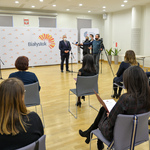 Jasne pomieszczenie. Prezydent Białegostoku mówi do mikrofonu. Obok stoi troje dziennikarzy. Kilka osób siedzi na krzesłach ustawionych w dużych odstępach.