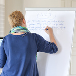 Kobieta z krótkimi blond włosami i kolorową chustą na szyi pisze tekst na kartce przyczepionej d tablicy. W drugiej dłoni trzyma włączonego laptopa.