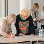 Blondynka z kremowym szalikiem oraz kobieta w bluzce z kolorową grafiką pochylają się nad kartkami leżącymi na stole. W tle stoją dwie rozmawiające kobiety.