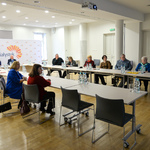 Przestronne pomieszczenie, grupa kilkunastu osób siedzi przy stole ustawionym w kształt litery U na środku pomieszczenia. 