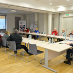 Przestronne pomieszczenie, grupa kilkunastu osób siedzi przy stole ustawionym w kształt litery U na środku pomieszczenia. Na ekranie rzutnika na jednej ze ścian wyświetlana jest prezentacja. 