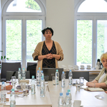 Prowadząca spotkanie kobieta o krótkich, ciemnych włosach stoi u szczytu stołu, przy którym siedzą członkowie spotkania.