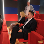 Zastępca Prezydenta Miasta Białegostoku oraz kilka innych osób siedzi na czerwonych fotelach w pomieszczeniu pogrążonym w półmroku. 