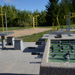 Siłownia zewnętrzna, na której są ustawione: na pierwszym planie stół do gry w piłkarzyki, dalej dwa stoły do gry w szachy wraz z siedziskami, a także urządzenia siłowni zewnętrznej.