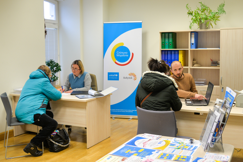 Punkt informacyjny. Pracownicy siedzą przy biurkach, rozmawiają z klientami punktu. W tle logo Centrum Spilno Unicef.