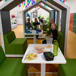Przestrzeń szkolna z małymi stolikami przy których jedzą dzieci. Nad częścią stolików zamontowane daszki w kształcie małych domków.