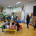 Duża otwarta przestrzeń szkolna. Na białych ścianach kolorowe rysunki zwierząt i roślin. Na jednej ze ścian mała ścianka wspinaczkowa i drabinki. Na środku pomieszczenia kolorowe stoliki i ławki przy których siedzą dzieci.