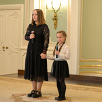 Dwie dziewczynki stoją na scenie trzymając w rękach mikrofony. Wyższa z dziewczynek trzyma za rękę młodszą. 