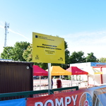 Na pierwszym planie żółta tablica informująca o realizacji projektu w ramach Budżetu Obywatelskiego 2024, za nią obok siebie namioty czerwone i żółte pod którymi znajdują się stanowiska dla seniorów