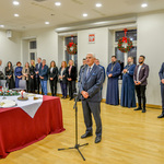 Prezydent Białegostoku mówi do mikrofonu stojąc obok udekorowanego stołu. W tle stojący przy ścianie goście specjalni.