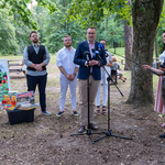 Przestrzeń parkowa wśród drzew. Uczestnicy konferencji prasowej stoją obok siebie. Zastępca prezydenta Białegostoku mówi do mikrofonu.